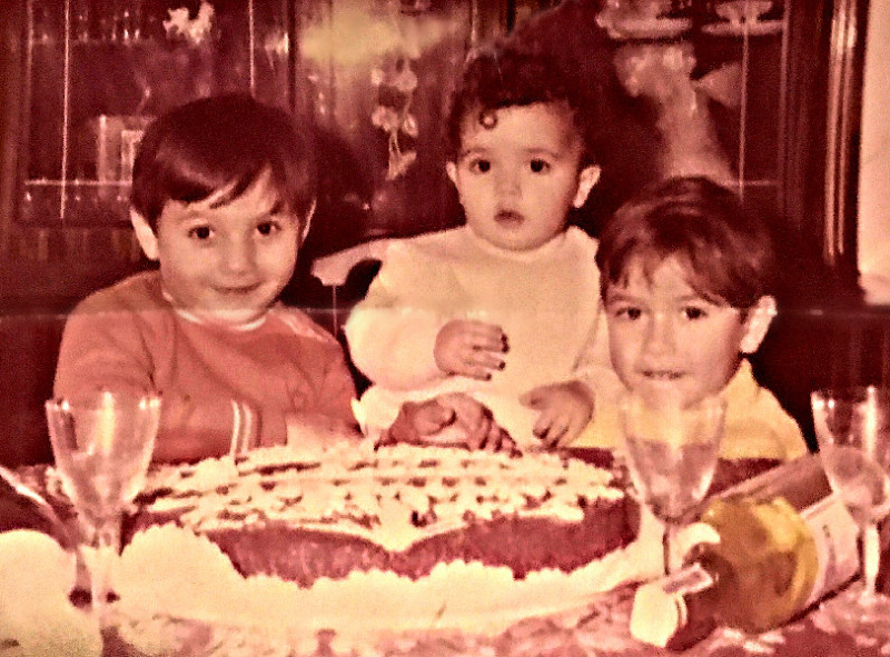 Colorized black and white photo of 3 small boys in front of a large cake.